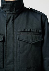 Schwarze Stoffjacke mit hohem Kragen, ausgestattet mit zwei Fronttaschen und sichtbaren Nahtdetails. Glatte Textur mit strukturiertem Design.