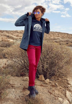 Draußenjacke in Dunkelblau, über einem marineblauen Shirt mit Bergdesign. Rote Hose, graue Schuhe und Spiegelsonnenbrille. Wüstenlandschaft im Hintergrund.