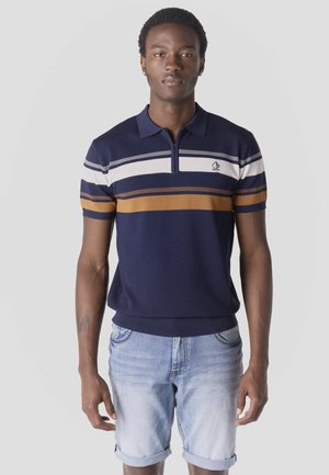 Joven con un polo azul marino con rayas blancas y marrones, combinado con pantalones cortos de mezclilla azul claro remangados, posando frente a un fondo liso.