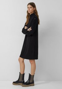 Zwarte gebreide jurk met hoge hals en knoopdetails op de schouder. Gepaard met zwarte leren enkellaarsjes en gestructureerde zijaccenten.