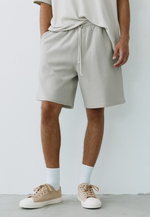 Uomo che indossa pantaloncini casual grigio chiaro, sneakers beige, calzini bianchi e una camicia larga abbinata, in piedi davanti a uno sfondo semplice.