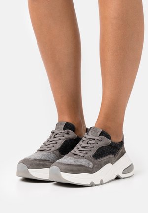 Nohy v šedých a černých semišových teniskách s bílými silnými podrážkami, stojící na bílém pozadí.