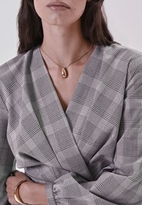 Top avvolgente a scacchi bianco e nero con maniche lunghe. Una collana con pendente a goccia dorata e un bracciale metallico lucido arricchiscono l'outfit.