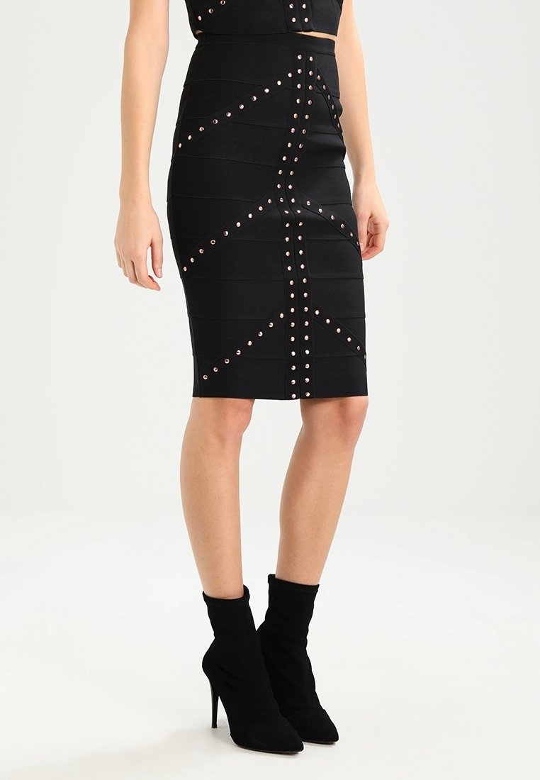 Femme portant une jupe noire au genou avec des motifs géométriques cloutés, associée à des bottines noires à talons hauts sur un fond uni.