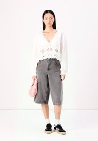 Haut en tricot ouvert blanc avec un motif floral en dentelle, associé à des culottes en jean gris taille haute. Le mannequin tient un sac rose clair et porte des baskets noires.