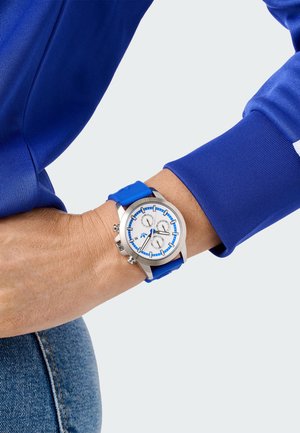 Montre avec bracelet en silicone bleu, boîtier en argent, cadran blanc, accents bleus et trois sous-cadrans. Poignet visible et tissu en denim.