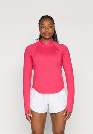 Maglietta sportiva rosa a maniche lunghe con colletto a mezza zip e orlo arrotondato, realizzata in materiale leggero. Indossata con pantaloni corti bianchi.