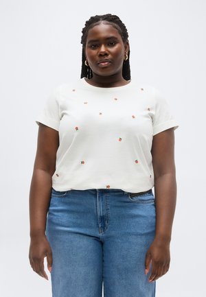 Donna nera con capelli intrecciati che indossa orecchini a cerchio dorati, una maglietta bianca con piccole stampe arancioni e jeans blu, in piedi contro uno sfondo semplice.