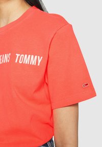 Korálové tričko s krátkým rukávem vyrobené z bavlny, s textem "TOMMY JEANS" v bílé barvě a malým logem značky v červené a bílé na rukávu.