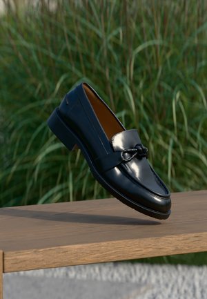 Mocassino nero lucido in pelle con dettaglio a nodo sulla cinghia, appoggiato su un bordo di una panchina di legno, con sullo sfondo erba alta e verde.