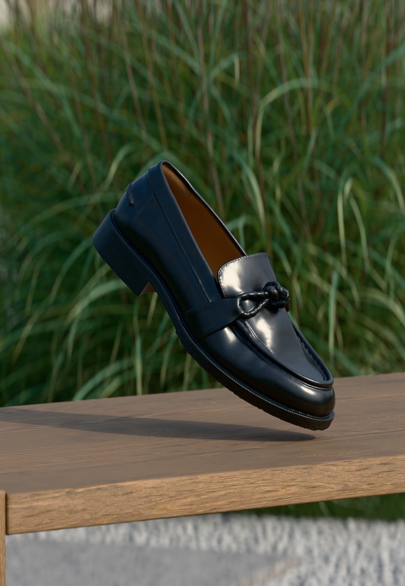 Λουστρινένιο μαύρο δερμάτινο μοκασίνι με λεπτομέρεια πλεξούδας στο λουράκι, ισορροπημένο σε μια ακμή ενός ξύλιν ου παγκάκι, με φόντο ψηλό πράσινο γρασίδι.