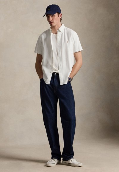 Fiatal férfi tengerészkék sapkában, fehér rövid ujjú gombos ingben, fehér póló fölött, sötét farmernadrágban, fehér sportcipőben, egyszínű bézs háttér előtt áll.