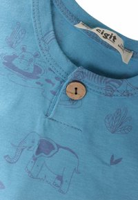 Blue cotton T-shirt with a round neckline, featuring a wooden button and an elephant pattern. Soft texture with subtle graphics in a lighter shade.