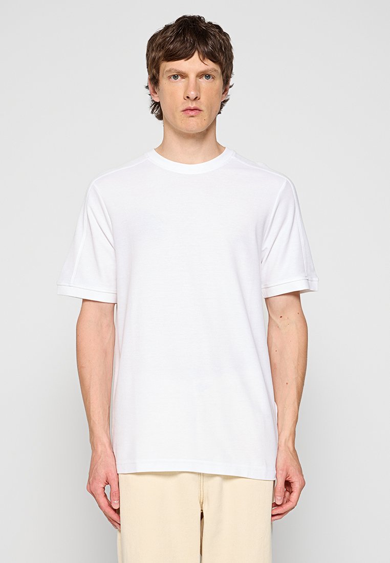 Junger Mann mit kurzem braunem Haar, der ein schlichtes weißes T-Shirt mit kurzen Ärmeln und eine hellbeige Hose trägt, steht vor einem grauen Hintergrund.