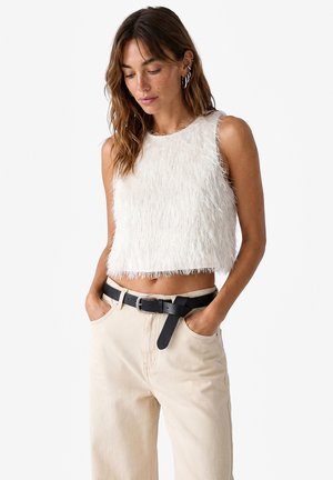 Donna che indossa un crop top bianco senza maniche e morbido, pantaloni beige a vita alta e cintura nera, mani nelle tasche, guarda verso il basso con un'espressione rilassata.