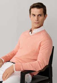 Mężczyzna siedzący na czarnym krześle, ubrany w jasnoróżowy sweter z dekoltem w serek nałożony na białą koszulę z kołnierzykiem oraz białe spodnie, patrzący w prawo.