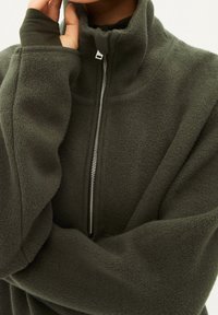 Pullover in pile verde scuro con colletto alto, cerniera frontale e fori per i pollici. Texture morbida e vestibilità rilassata. Design minimalista senza fantasie.