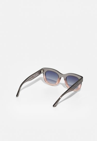 Lunettes de soleil avec une monture transparente grise et rose, des verres dégradés bleus, et un design élégant et angulaire avec des branches fines.