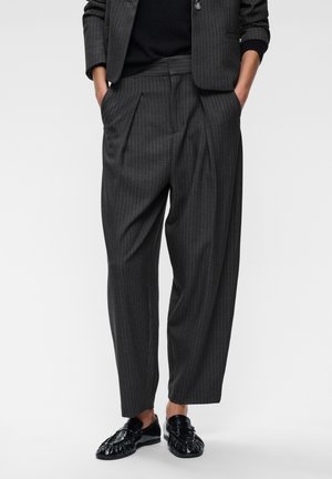 Vue recadrée d’une personne portant un pantalon à pinces anthracite à fines rayures, un blazer assorti et des mocassins noirs, les mains dans les poches.