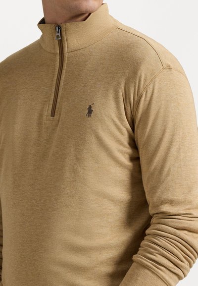 Kreminės spalvos pusiau užtraukiamas megztinis, pagamintas iš minkštos, tekstūruotos medžiagos, su mažomis tamsiomis logotipų ant kairiojo krūtinės ir raukšlėtomis rankogalių dalimis.