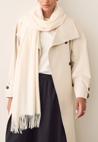 Beige trench coat with large lapels and black buttons, paired with a cream scarf featuring fringed edges. Black pants are also visible.