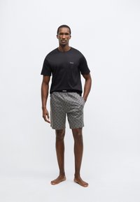 Μαύρο βαμβακερό t-shirt με λογότυπο, συνδυασμένο με γκρι σορτς που έχουν επαναλαμβανόμενο μαύρο σχέδιο και ελαστική μέση. Το μοντέλο στέκεται ξυπόλυτο.