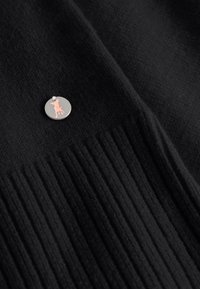 Zwarte stof met verticale ribbelstructuur, met aan de linkerkant een ronde metalen pin met een roze silhouet.