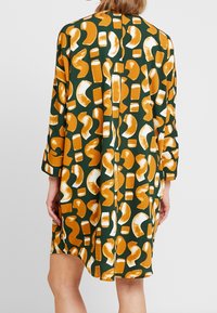 Grön klänning med orange och vita abstrakta mönster, långa ärmar och avslappnad passform. Tyget verkar mjukt med en lätt glans.