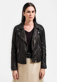Veste de moto en cuir noir avec des zips argentés, devant asymétrique et épaulettes. Associée à un haut noir texturé et une jupe légère.