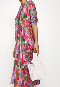 Farbiger Blumenkleid aus pinkem, grünem und rotem Stoff mit langen Ärmeln; kombiniert mit einer hellpinkfarbenen, strukturierten Mini-Tasche mit Schleifendetail.
