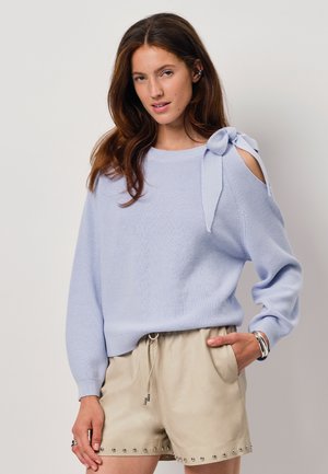 Lichtblauwe gebreide trui met open schouderontwerp en strikdetail. Comfortabele pasvorm met lange mouwen, gecombineerd met beige shorts met studs.