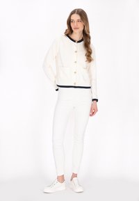 White cotton blend cardigan with navy trim and gold buttons, featuring two front pockets. Paired with white fitted jeans and sneakers.