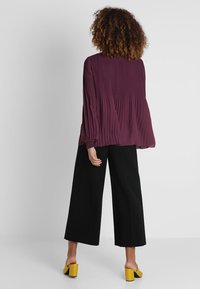 Kvinna med lockigt hår klädd i en plisserad plommonblus, svarta vida byxor och gula klackskor med blockhäl, stående med ryggen mot kameran.