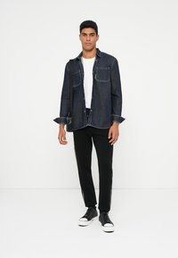 Dunkelblaue Jeanshemd mit zwei Brusttaschen, geknöpfter Front, kombiniert mit schwarzen Hosen mit weißen Seitestreifen und schwarzen Sneakers.