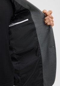 Innenfutter eines grauen Blazers mit Muster und glattem schwarzem Stoff; verfügt über eine Reißverschlusstasche und kontrastierende Nähte, die ein strukturiertes Design zeigen.