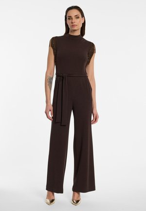 Femme portant une combinaison sans manches marron foncé avec des franges perlées aux épaules, une ceinture à la taille, jambes larges, et des chaussures dorées à bouts pointus.