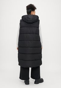 Melnā pūkainā vestē ar kapuci, kas izceļas ar quilted dizainu un slāņveida izskatu. Modelis to valkā virs pelēka džempera un melnām biksēm.