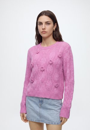 Femme aux longs cheveux bruns portant des boucles d'oreilles créoles en or, un pull en maille texturée rose avec des détails floraux, et une jupe en denim bleu clair.
