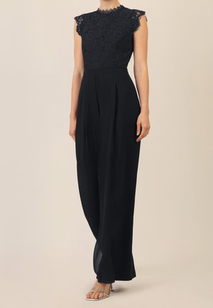 Combinaison noire sans manches avec corsage en dentelle, col montant, pantalon pattes d'éléphant, et talons argentés à bout ouvert sur fond beige.