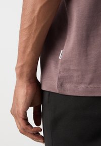 T-shirt mauve cu mâneci scurte, dintr-un material texturat și cu un etichetă pe lateral, purtat cu pantaloni negri, arătând o mână odihnindu-se pe lateral.
