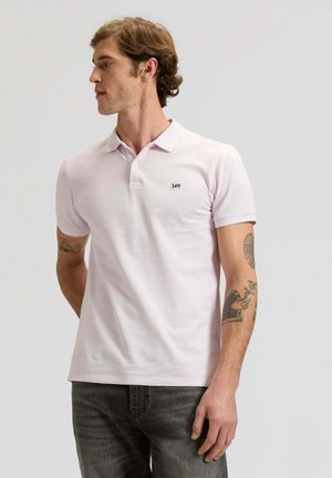 Mann mit Tätowierungen, der ein hellrosa Lee Poloshirt und dunkle Jeans trägt, schaut nach links vor einem einfarbigen hellen Hintergrund.