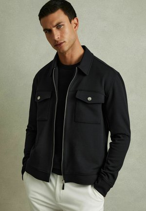 Veste noire zippée avec deux poches avant, fabriquée en tissu lisse. Dotée d'un col et de poignets côtelés, portée sur un top noir et un pantalon blanc.