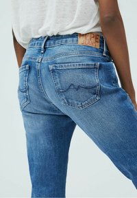 Jeans de mezclilla azul con una cintura de altura media, que presentan una etiqueta de cuero marrón en la parte trasera y dos bolsillos traseros con sutiles detalles de costura.