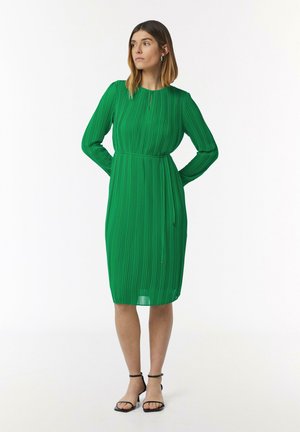 Robe plissée verte avec des manches longues, un col rond et une taille cintrée. Texture lisse, ourlet au niveau du genou, assortie à des sandales noires à bride cheville.