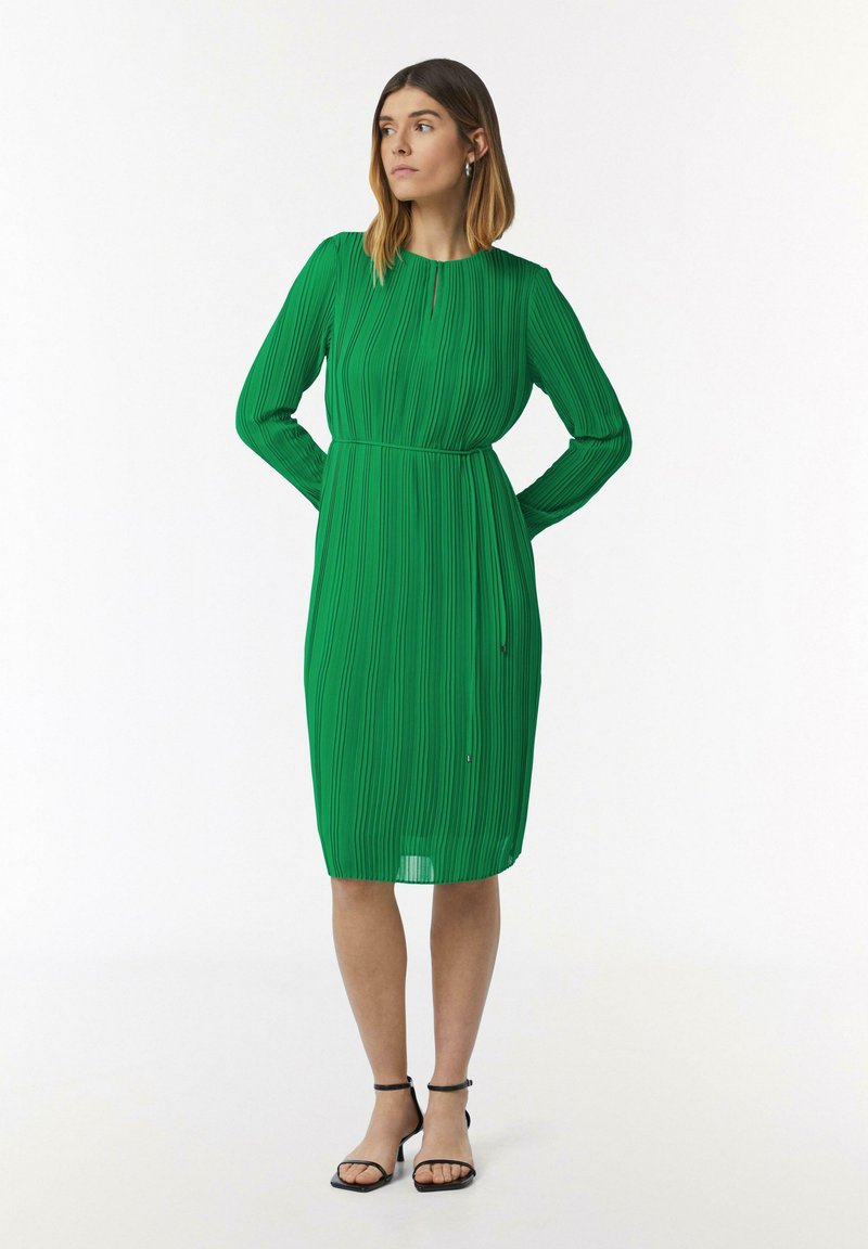 Robe plissée verte avec des manches longues, un col rond et une taille cintrée. Texture lisse, ourlet au niveau du genou, assortie à des sandales noires à bride cheville.