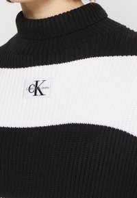 Pull en maille rayé noir et blanc avec un col montant côtelé et un patch logo Calvin Klein Jeans sur le devant. Tissu texturé.