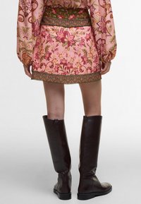 Floral-patterned pink skirt with green waistband and ornate trim; paired with knee-high, glossy brown leather boots.