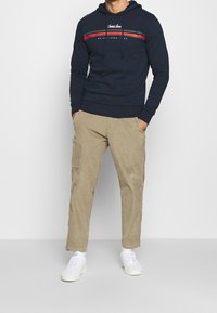 Tmavě modrá mikina s kapucí a šňůrkami, s logem, doplněná světle hnědými cargo kalhotami. Doplňky zahrnují bílé tenisky s texturovaným designem.