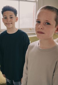 To drenge står tæt sammen. Den ene har en tekstureret sort langærmet trøje på, mens den anden har en lysebeige rundhalset sweater.