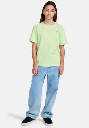 T-shirt en coton vert clair à manches courtes, associé à un jean bleu ample et des baskets noires. Design simple sans motifs ni logos visibles.